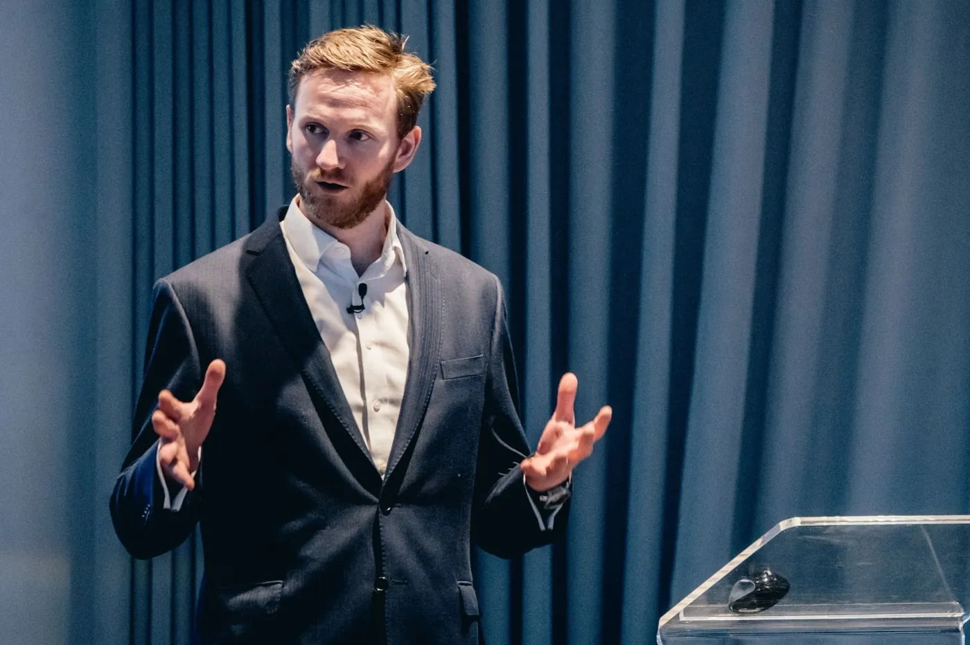Mark Agnew delivering a motivational keynote on stage in a dark suit, gesturing to a corporate audience